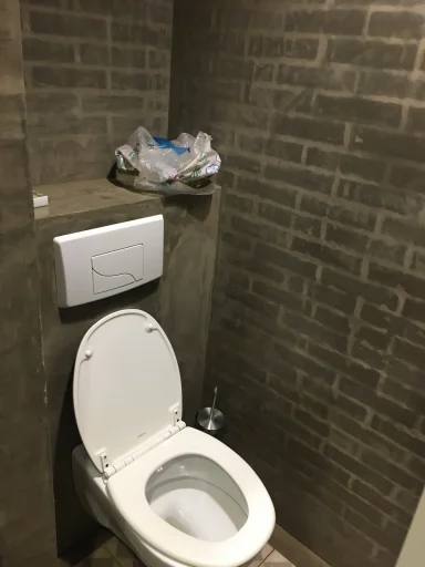 Avant, wc Toilette blanche avec mur en briques grises, un sac en plastique sur un rebord.